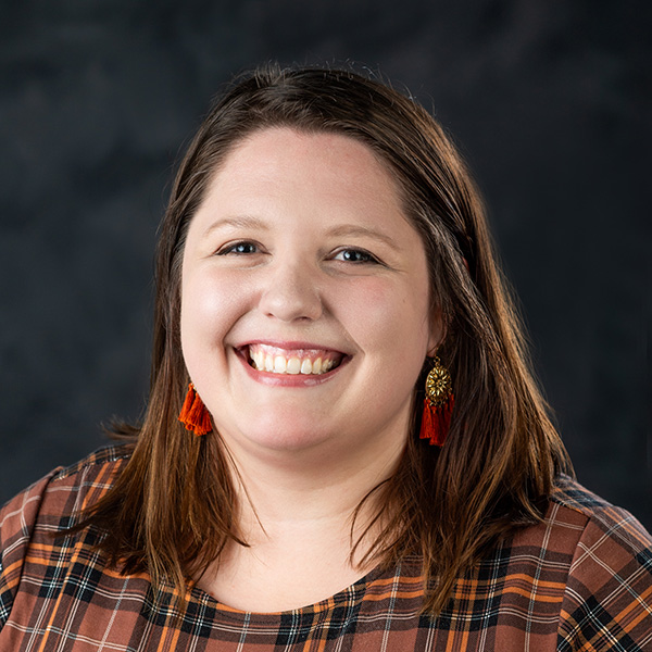 A person with shoulder-length brown hair, wearing a plaid shirt and red earrings, smiles against a dark background.