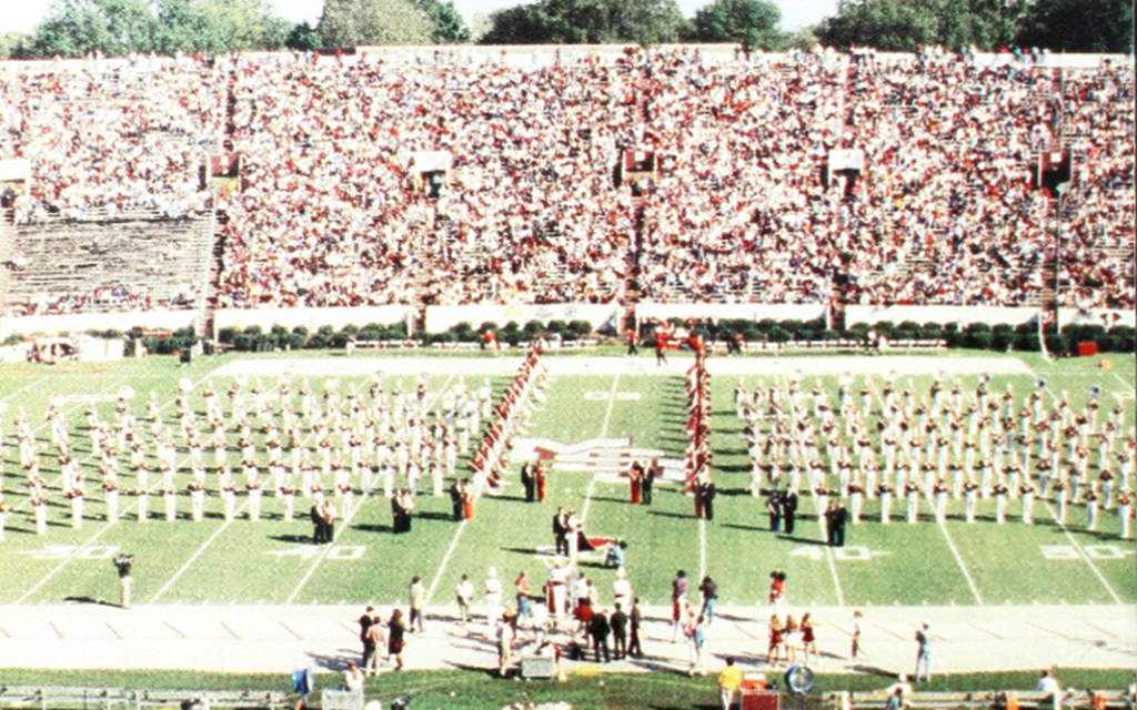 A marching band performs on a football field, forming a design. A large crowd fills the stadium seats in the background.