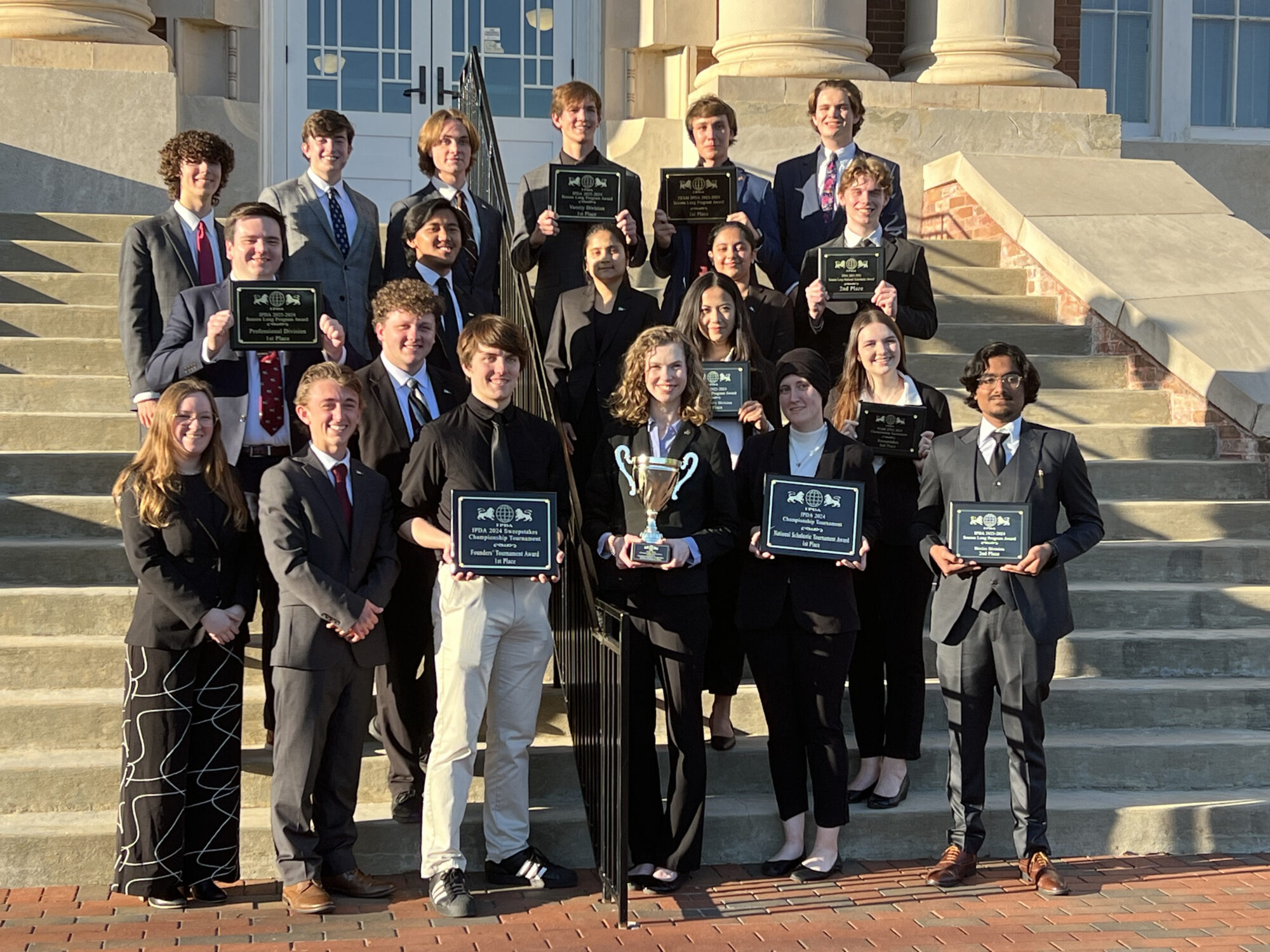 A group of people stands on steps in formal attire, holding plaques and a trophy.