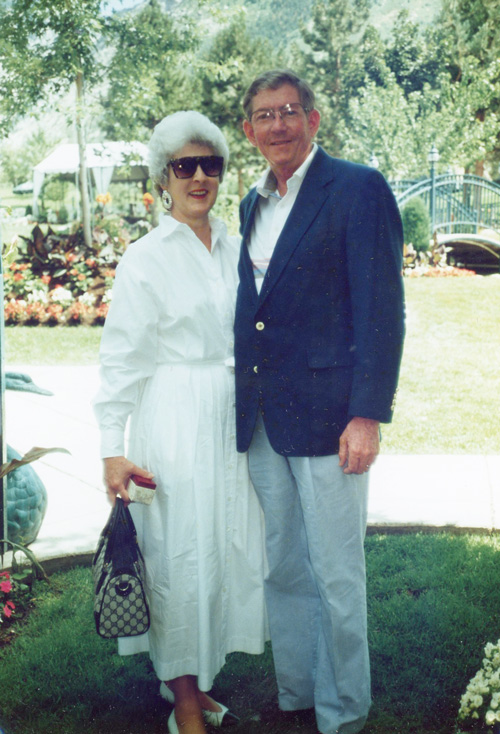 A couple stands together outdoors, the woman wearing a white dress and sunglasses, and the man in a dark blazer. They are on a grassy area with trees and a bridge in the background.