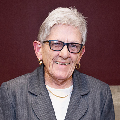 An elderly person with short white hair and glasses smiles while wearing a striped suit jacket and gold hoop earrings.