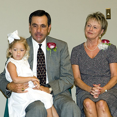 A man in a suit holds a young girl in a white dress with a bow. A woman in a patterned dress sits beside them. Both adults wear red rose corsages.