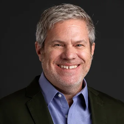 A person with short gray hair smiles, wearing a blue shirt and dark jacket against a dark background.