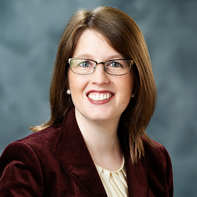 A person wearing glasses, a blazer, and a shirt is smiling in a portrait set against a gray background.