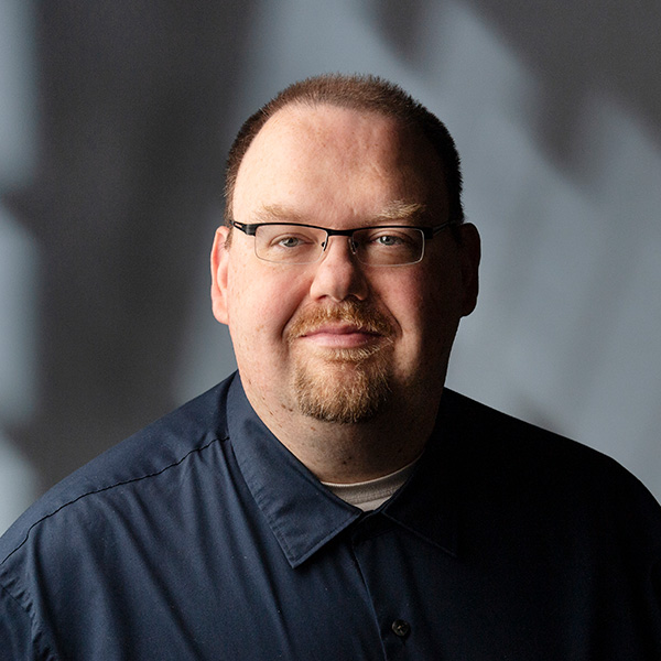 A man with glasses and a trimmed beard wearing a dark shirt, standing against a softly lit background.