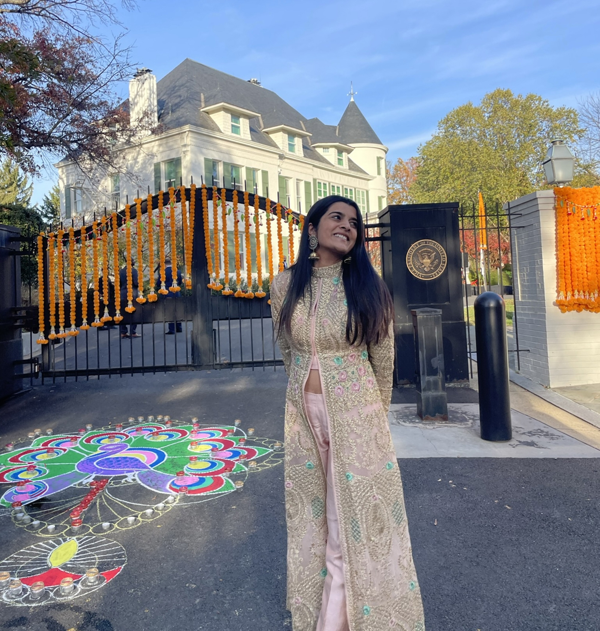 A woman in traditional attire stands in front of a decorated gate with a colorful rangoli on the ground. A large house is visible in the background.