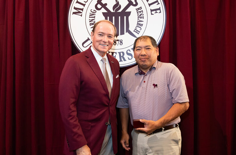 Two individuals stand in front of a maroon curtain with a university emblem. One person holds an award.