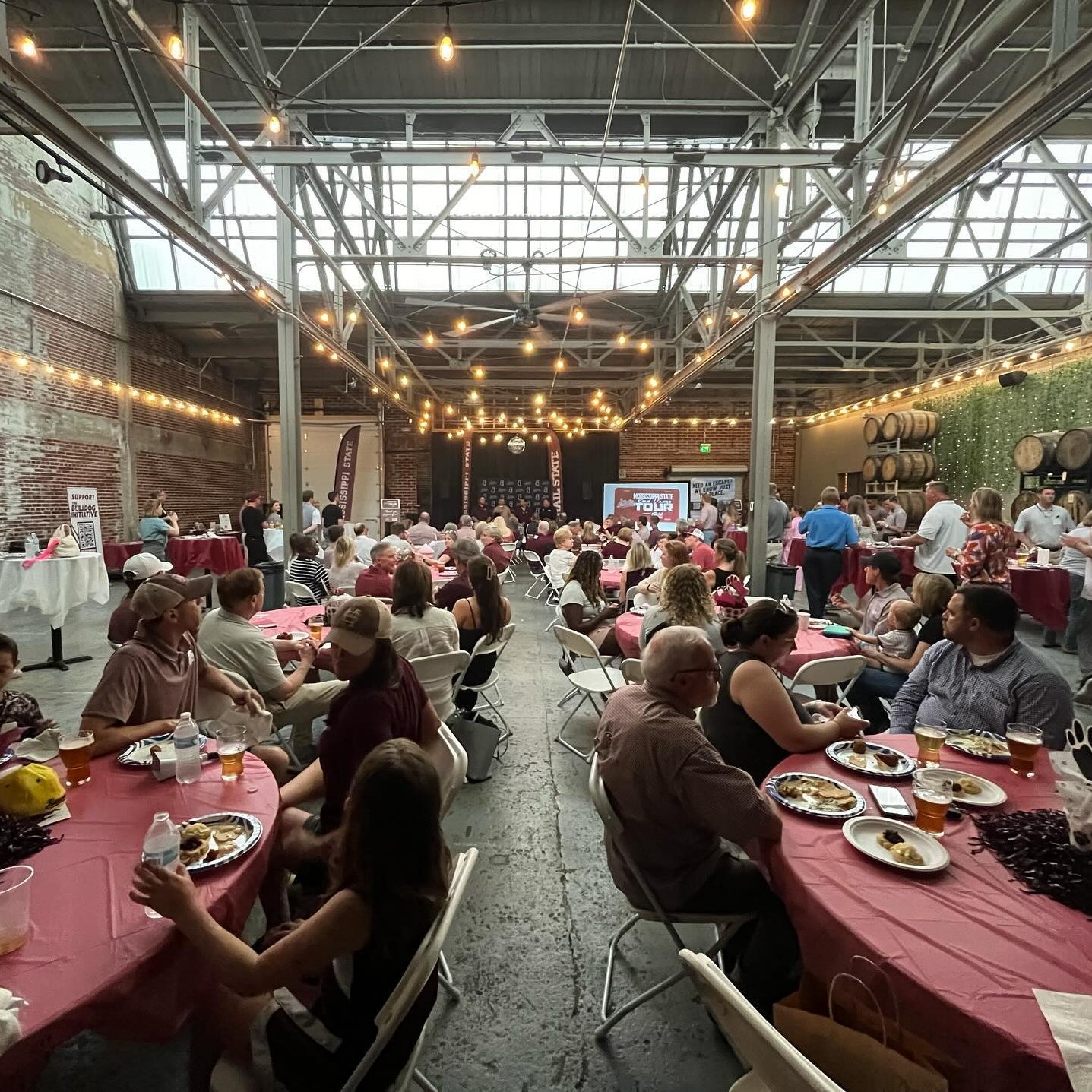 People seated at tables in a large, industrial-style venue with string lights. A stage is visible in the background with a presentation screen.