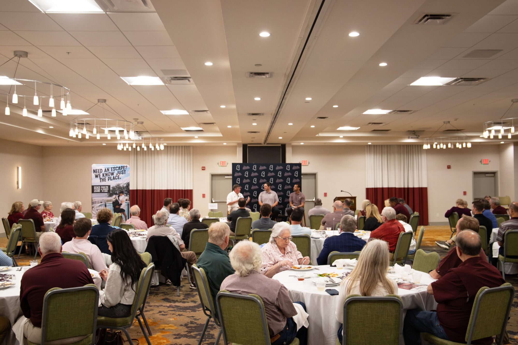 A group of people seated at tables in a conference room are listening to a speaker at the front. A large banner and a poster are displayed behind the speaker.