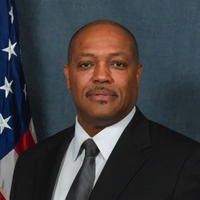 Man in a suit poses in front of a U.S. flag with a neutral expression.