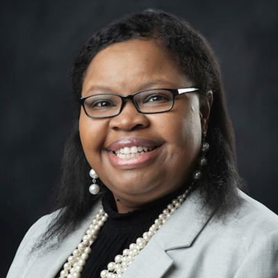 A person with glasses and a wide smile is wearing a gray blazer, black top, and pearl necklace against a dark background.