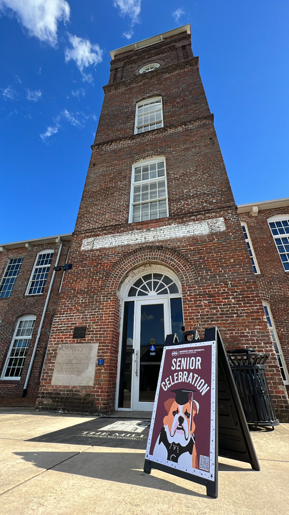 Tall brick building with arched entrance under a clear blue sky. Signboard in front reads "Senior Celebration" with an illustration of a dog wearing sunglasses.