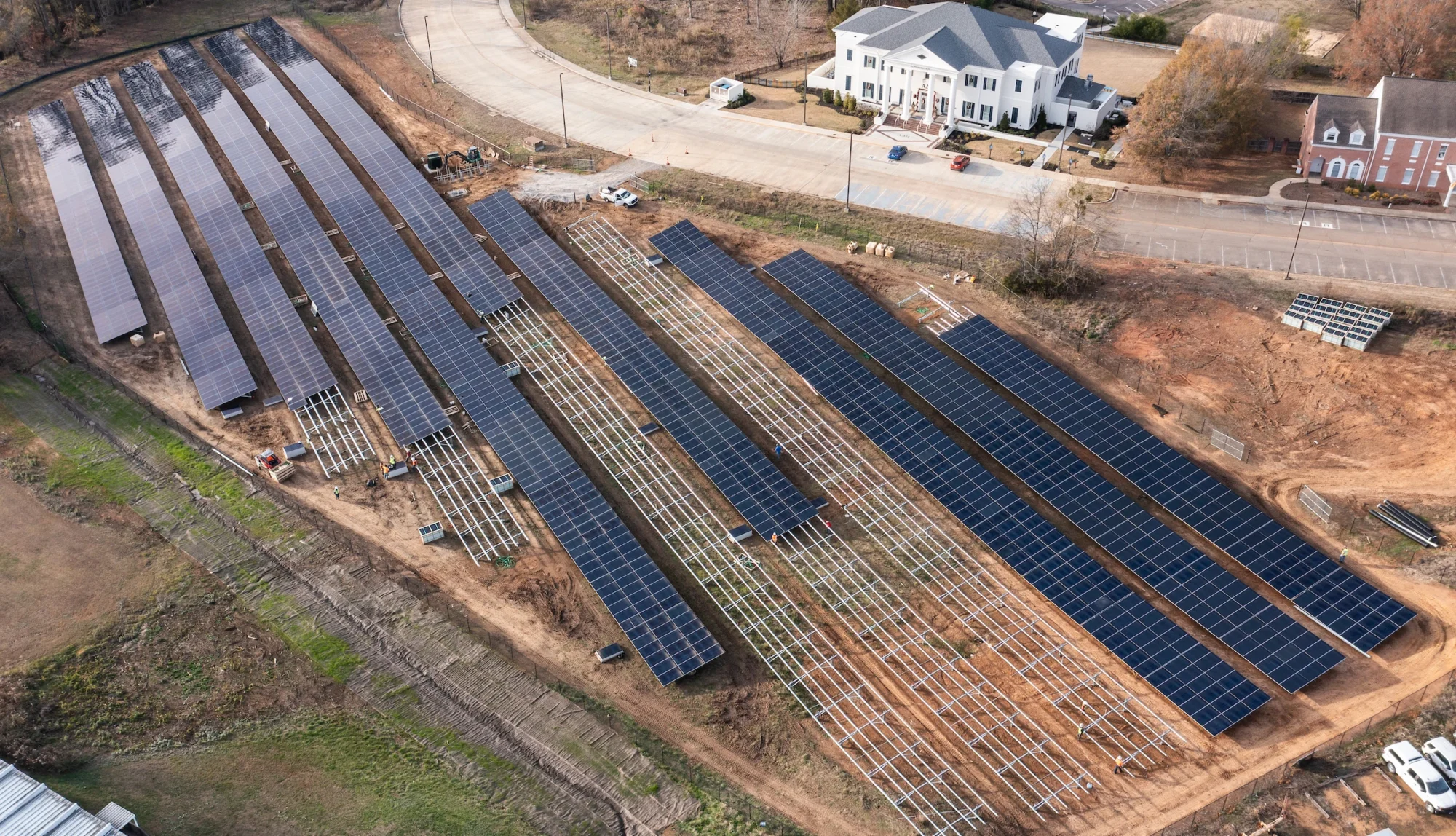 Aerial view of a solar panel installation near residential houses, showcasing rows of panels on brown soil.