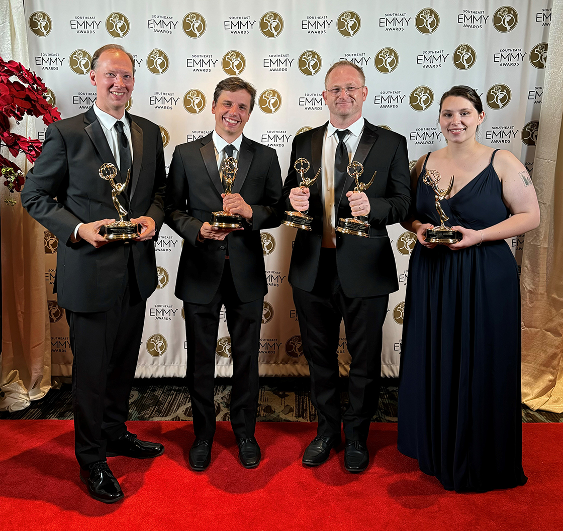 Four people in formal attire stand on a red carpet, each holding an Emmy award, in front of an Emmy-themed backdrop.