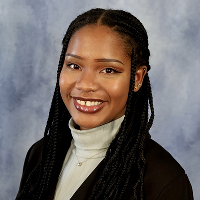 Portrait of a person with long braided hair, smiling, wearing a black blazer over a light gray turtleneck, set against a blue-gray background.