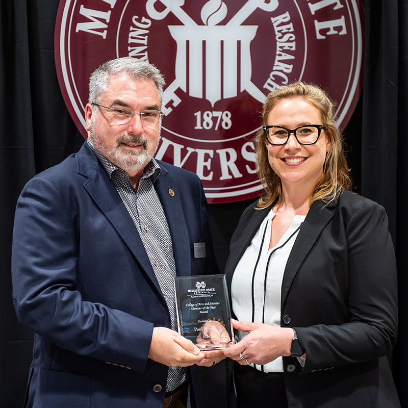 Two people in formal attire holding an award together in front of a university emblem.