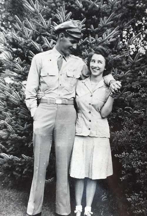 A man in a military uniform stands beside a woman in a dress. They are smiling at each other, with a large tree in the background.