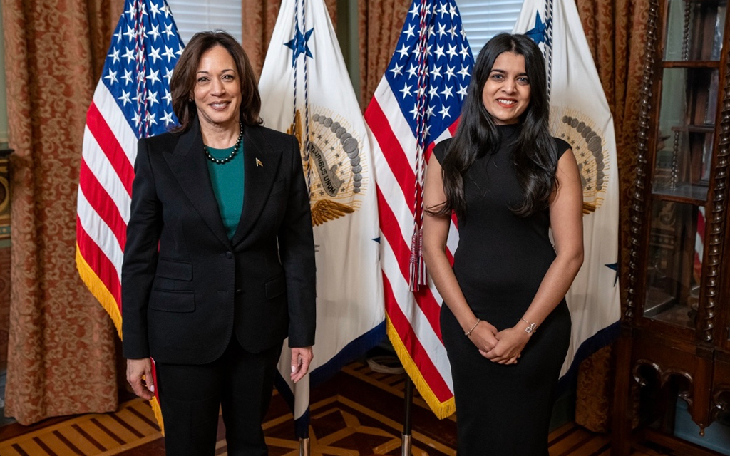 Two women stand indoors in front of U.S. flags, both wearing formal attire.