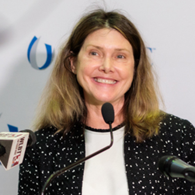 A woman smiling while standing at a podium with microphones, wearing a black and white outfit.