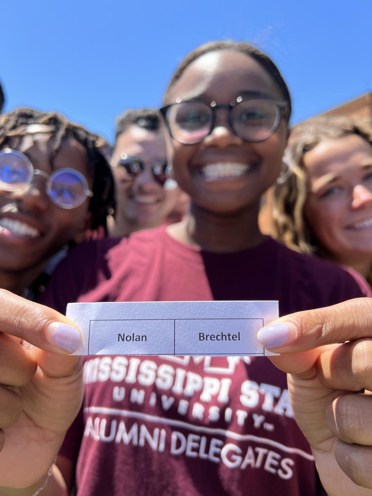 A group of smiling people, one in a "Mississippi State University Alumni Delegates" shirt, holding a paper with "Nolan Brechtel" written on it.