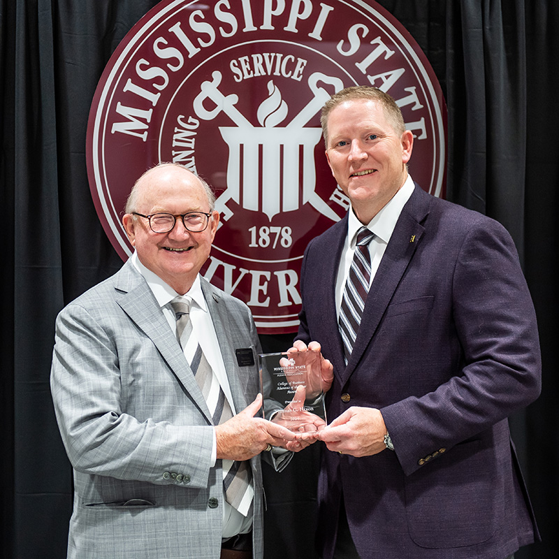 Two men in suits stand in front of a Mississippi State University emblem, smiling and holding an award together.