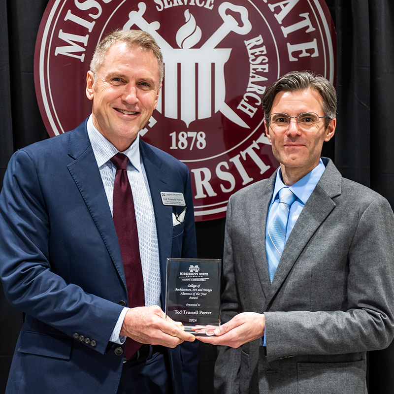 Two men in suits holding an award plaque in front of a Missouri State University banner.