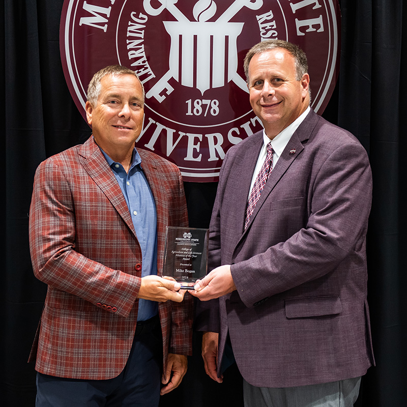 Two men in suits stand in front of a university banner; one is presenting an award to the other.