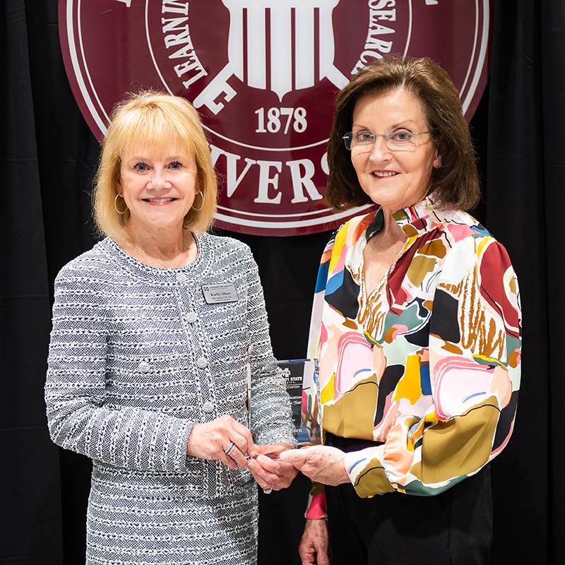 Two women stand smiling, one holding an award. They are in front of a maroon backdrop with the year 1878 and the word "University.