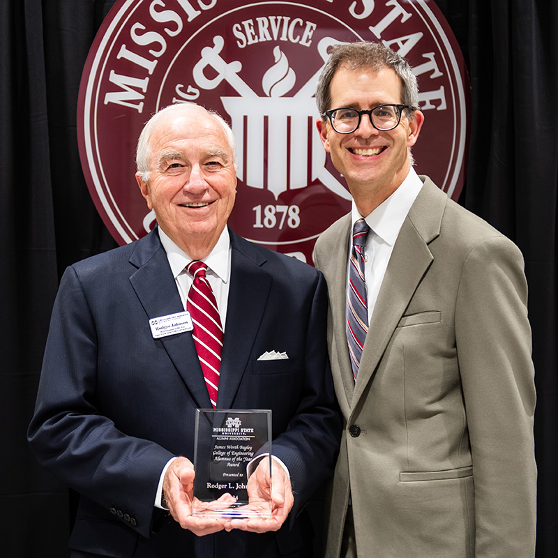 Two men in suits smiling, one holding an award. They stand in front of a Mississippi State University sign.