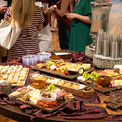 A table featuring a charcuterie board with assorted cheeses, meats, fruits, and pastries. People are standing in the background near a drink dispenser.