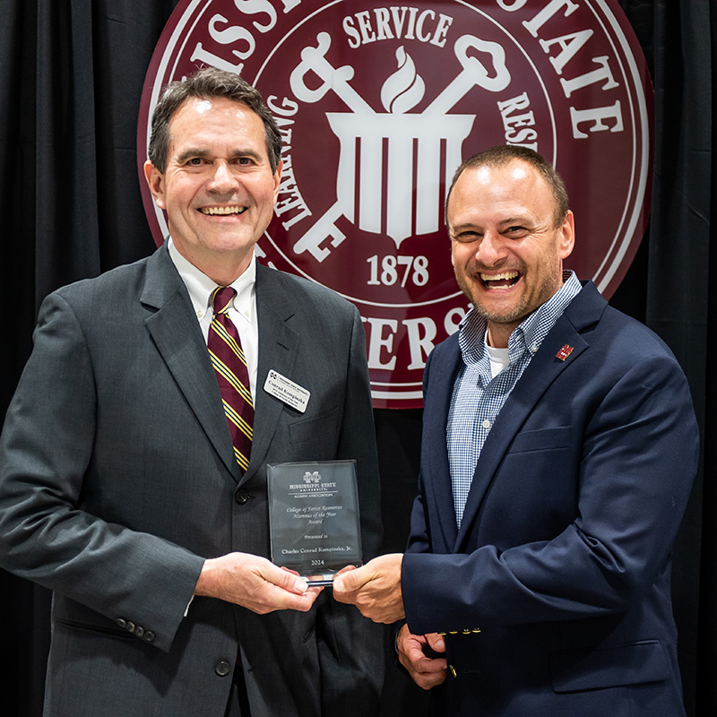 Two men in suits stand in front of a Mississippi State University seal, holding a glass award together and smiling at the camera.