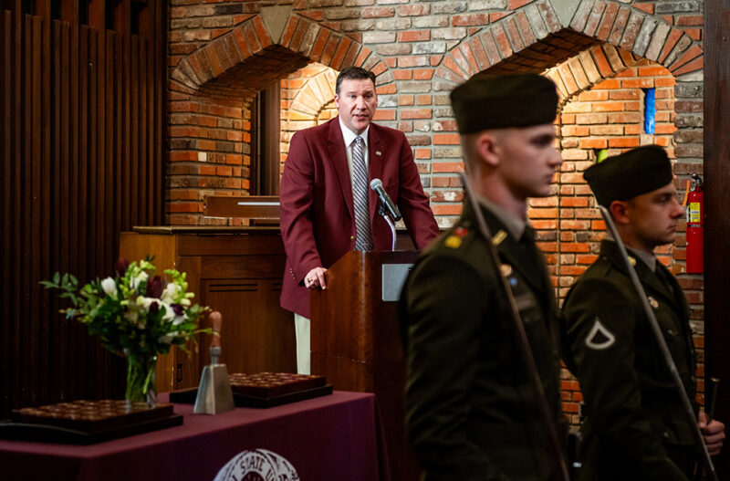 A man in a maroon jacket speaks at a podium. Two uniformed individuals stand in the foreground.