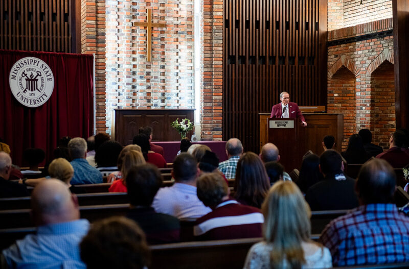 Speaker at a podium in a chapel with an audience seated, featuring a cross, brick walls, and a Mississippi State University banner.