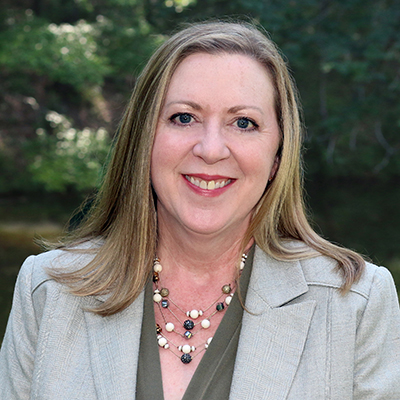 A woman with long hair smiling, wearing a light-colored blazer and a beaded necklace, in an outdoor setting with greenery in the background.