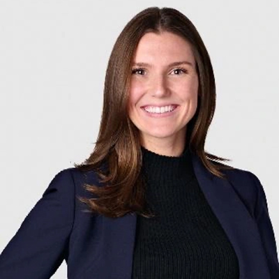 A person with long brown hair, wearing a black top and a blazer, smiles against a neutral background.