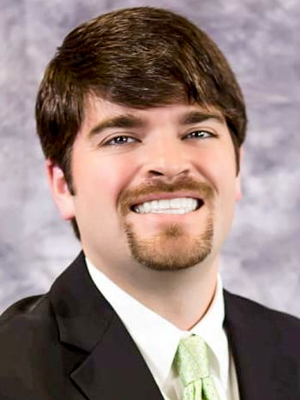 Smiling man in a suit with a light green tie against a gray background.