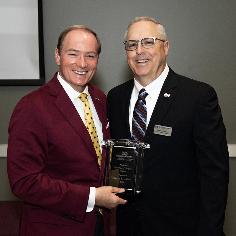 Two men in suits smiling while holding an award in an indoor setting.