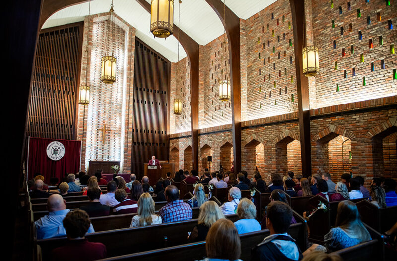 People seated in a church service, facing a speaker at a podium in a brick-walled chapel with stained glass windows.