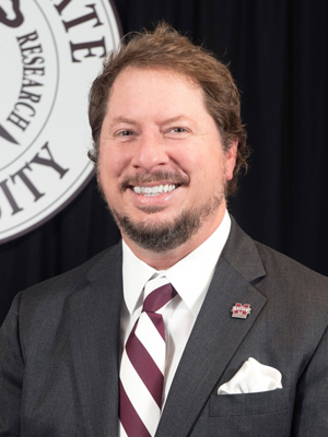 A man in a suit and striped tie is smiling in front of a logo backdrop with a maroon and white motif.