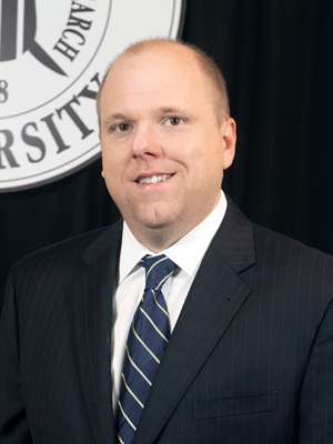 A man in a suit and tie stands in front of a university logo.