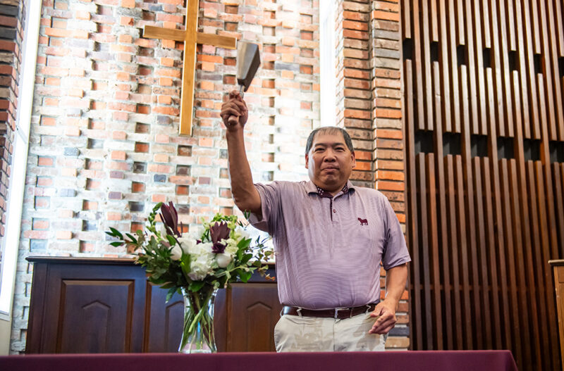 A person raises an instrument inside a church with flowers on the table, and a wooden cross on the brick wall behind them.