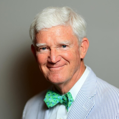 A smiling older man with white hair wears a light striped suit and a green patterned bow tie, standing against a plain background.