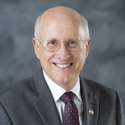 A smiling man wearing glasses and a suit stands in front of a gray background.