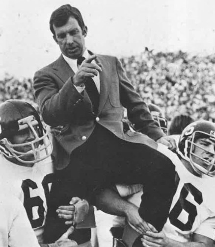 A man in a suit is being lifted by football players on their shoulders during a celebratory moment on the field.