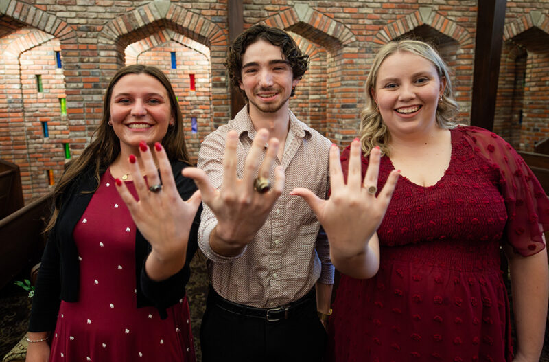 Three people smiling and showing rings on their outstretched hands in front of a brick wall with arched window designs.