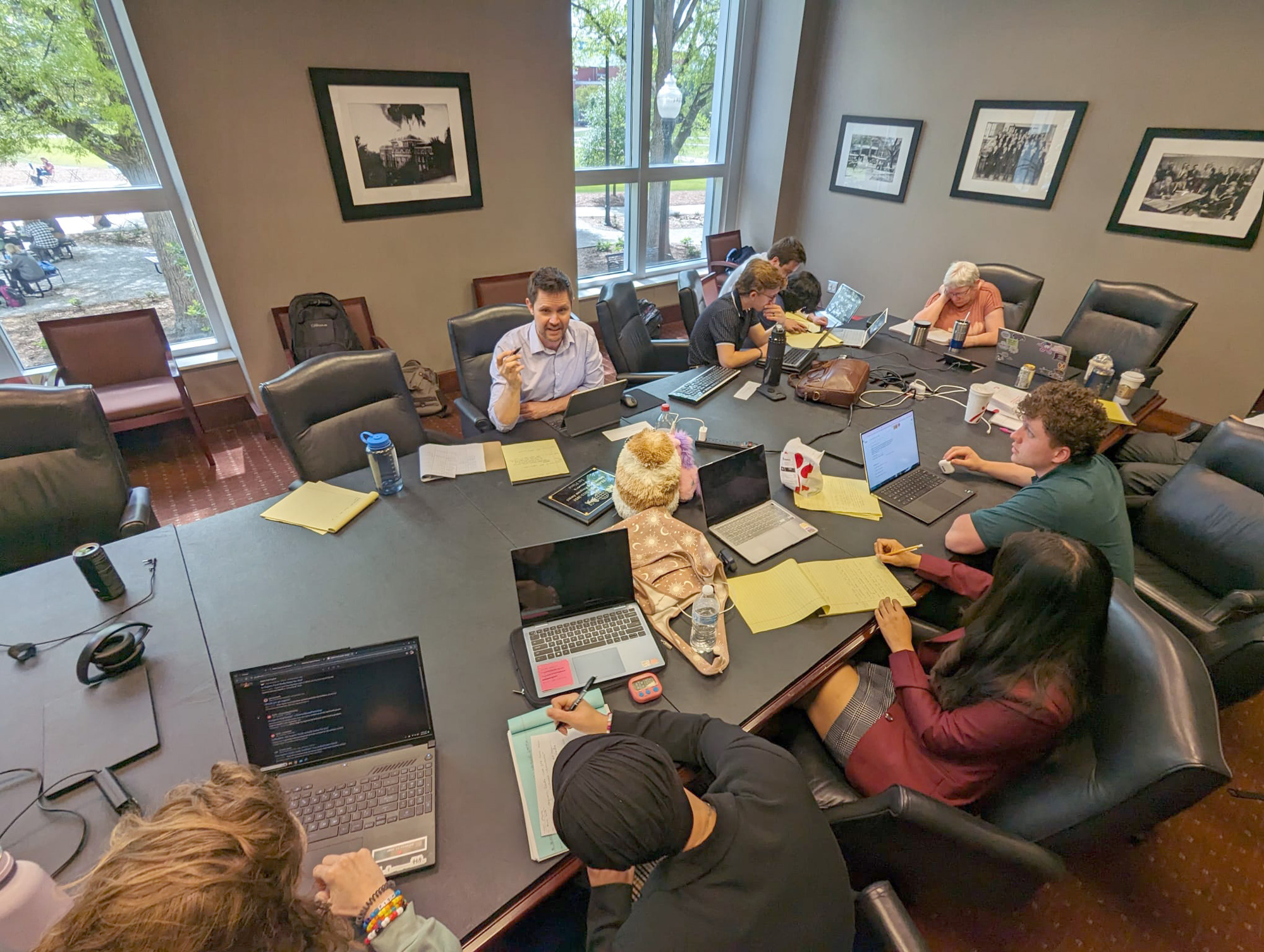 A group of people in a meeting room sit around a large table with laptops, notebooks, and drinks. Some are taking notes while others are engaged in conversation. Framed pictures are on the walls.