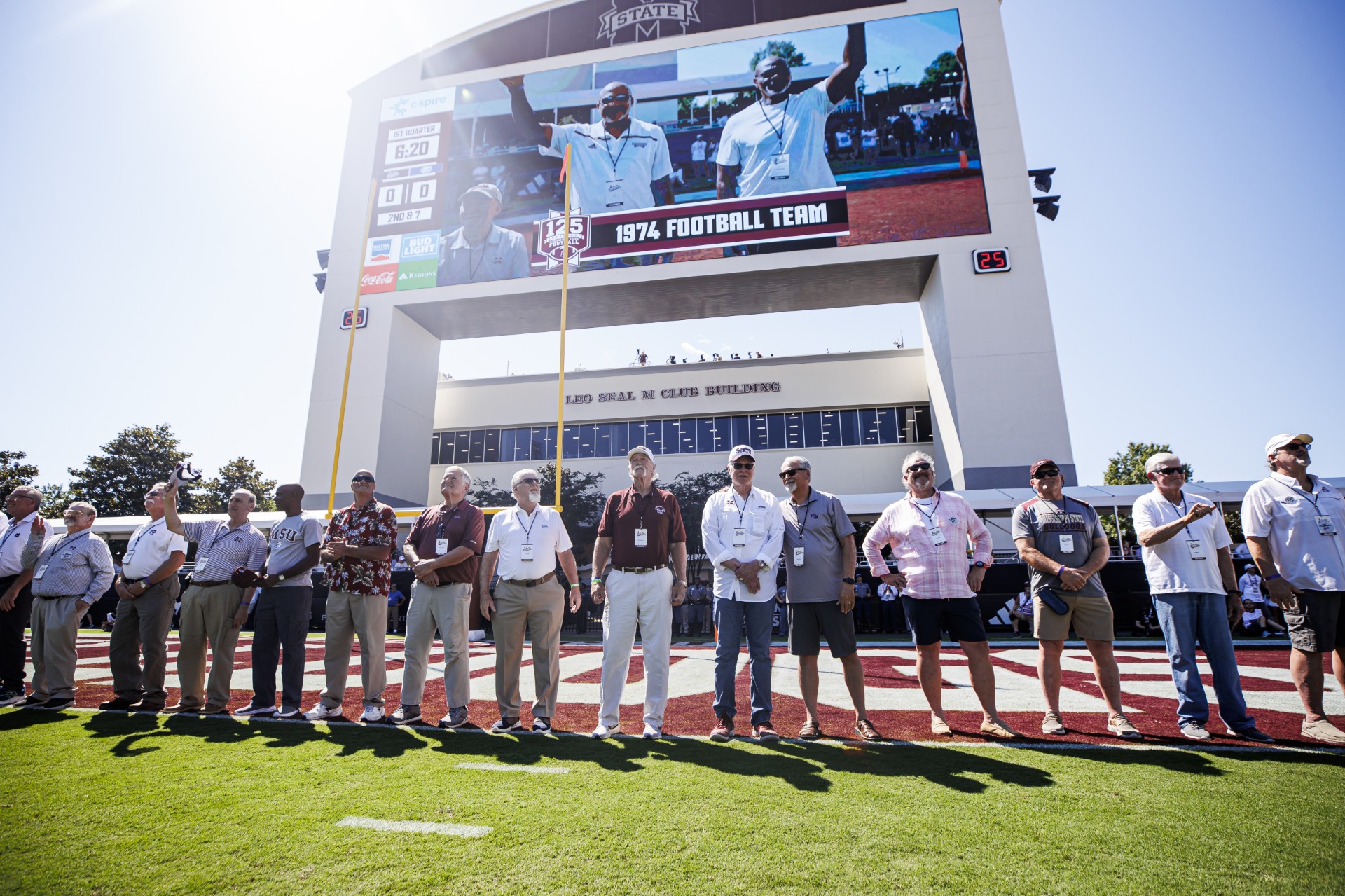 A group of people stands on a football field, posing for a photo in front of a scoreboard displaying "1974 Football Team.