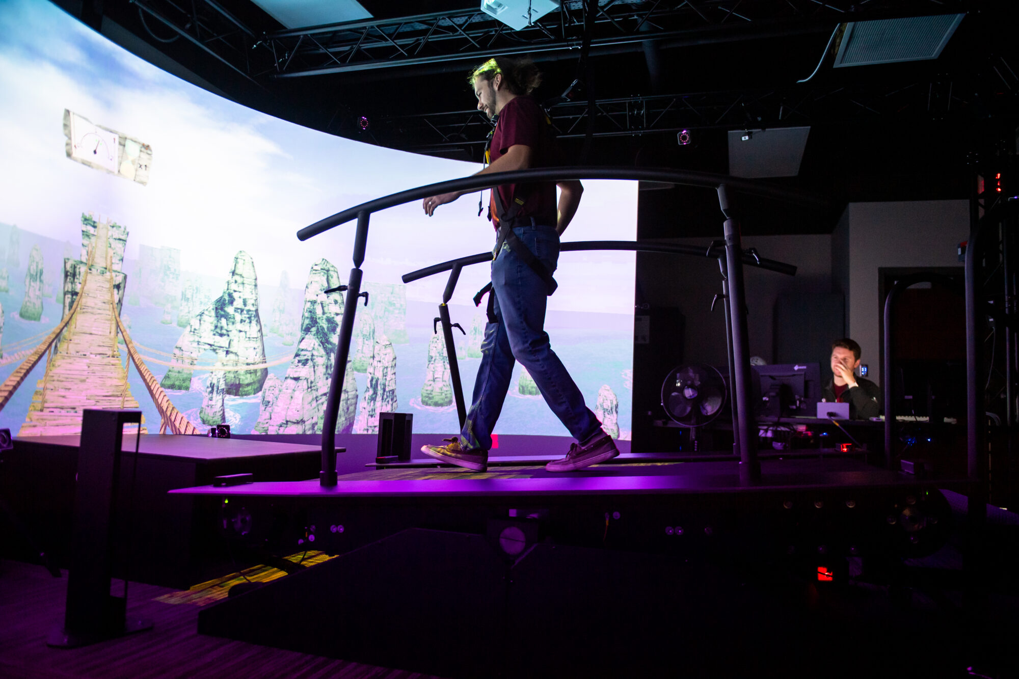 Person walking on a treadmill in a virtual reality setting with colorful landscape projection and a seated technician nearby.