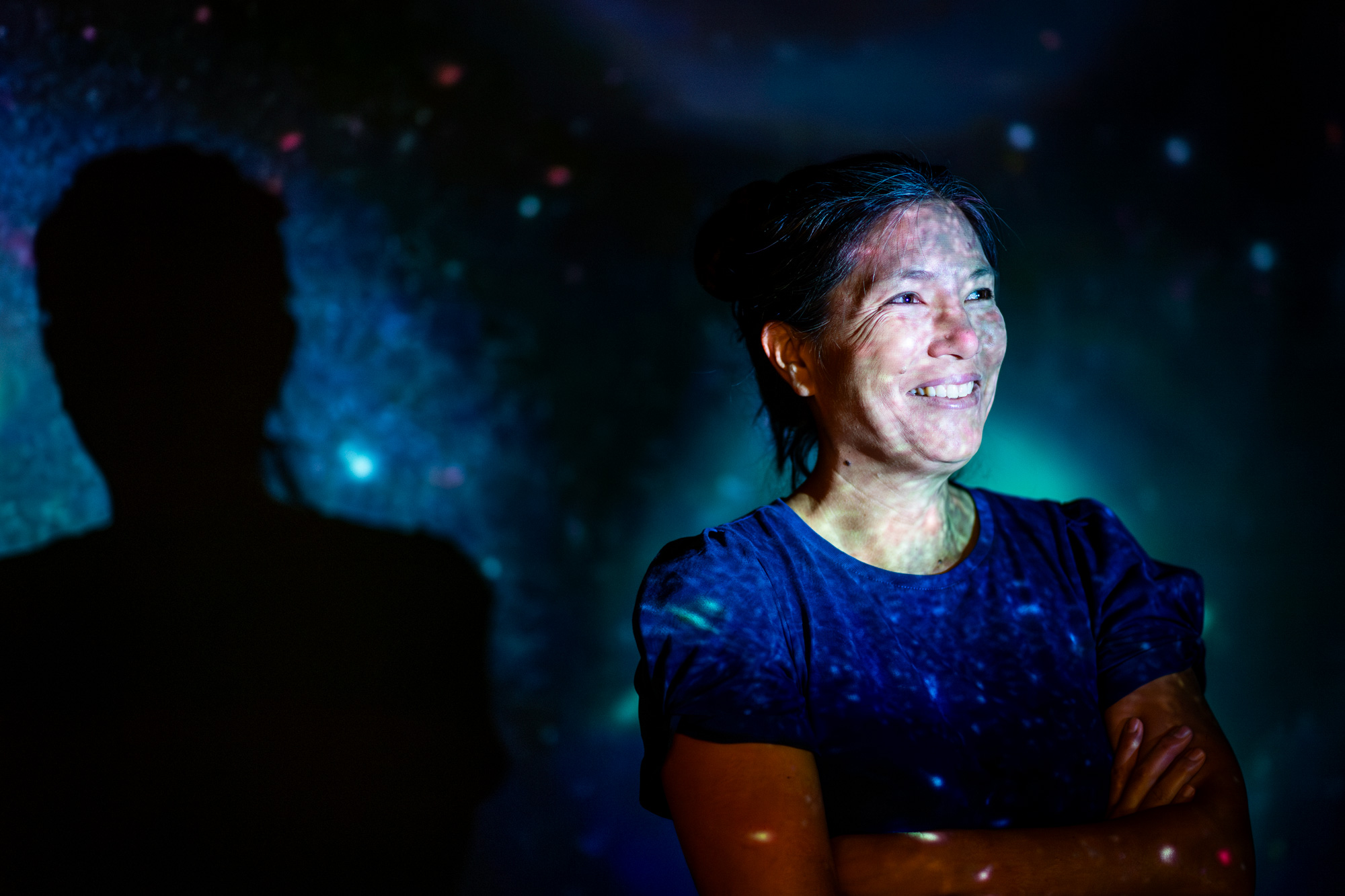 A person stands smiling with arms crossed against a dark backdrop with colorful lights, creating a starry effect.