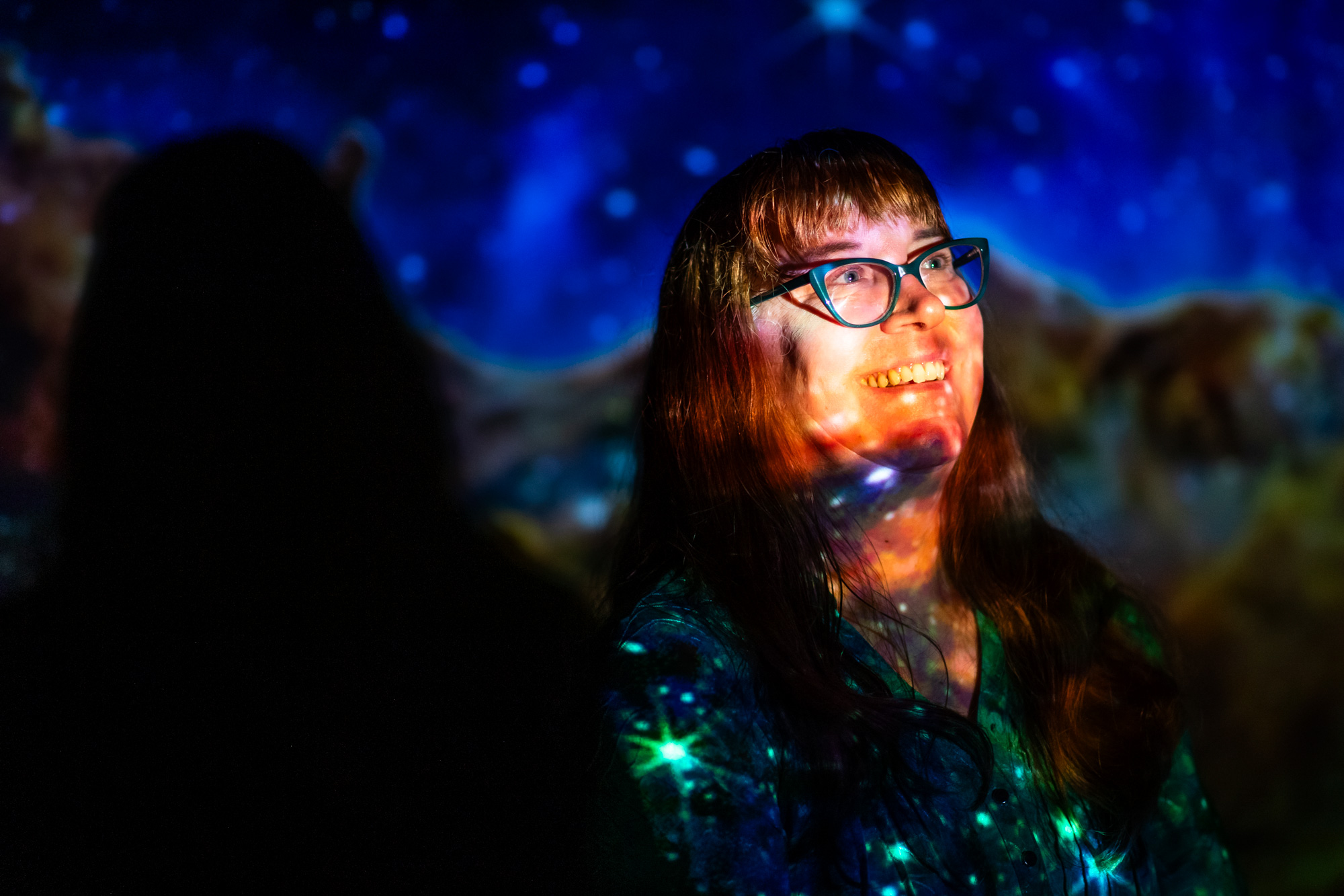 A person with glasses smiles under colorful, star-like projections against a dark background.